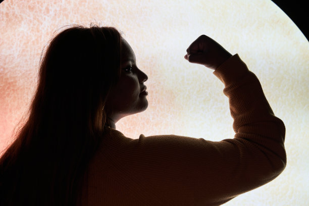 一个富有魅力和自信的年轻非洲裔美国女性的剪影，飘逸的头发和弯曲的身体，在工作室的墙壁前摆出独立的姿势，展示她的力量和肌肉，绷紧她的二头肌。图片下载