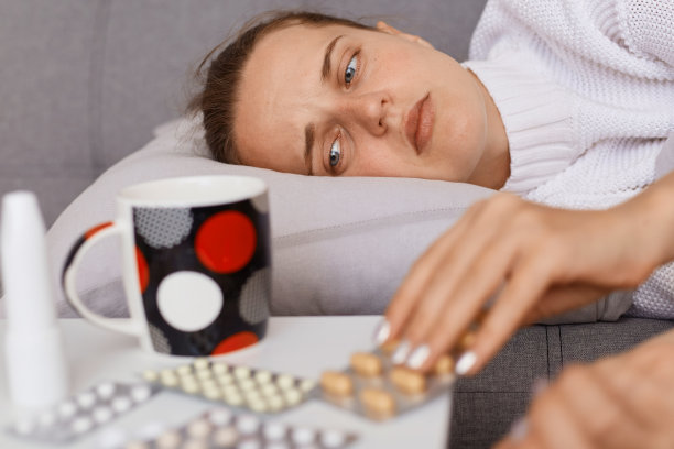 在室内拍摄的照片中，一名年轻女子躺在沙发上吃药，看着药片和茶杯或水，表现出了流感症状，表现出悲伤的情绪和悲伤。图片下载