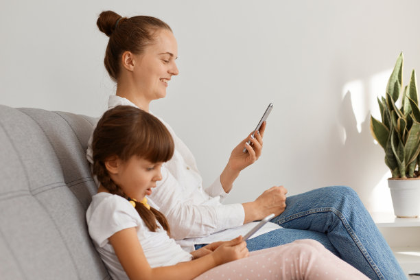 照片中的美女穿着白衬衫和牛仔裤，和女儿坐在沙发上，通过手机上网，在社交网络上观看有趣的视频或朋友的照片。图片下载