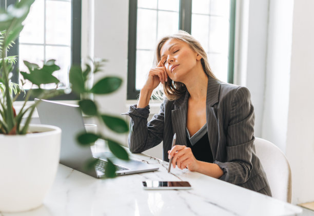 年轻疲惫的金发女子，一头长发，穿着时髦的灰色西装，在明亮的办公室里用笔记本电脑工作图片下载