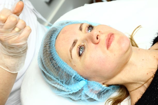 美容院。一位戴着医用手套和防护面罩的美容师正在为客户的脸颊做水螅脱皮手术。侧视图。关闭了。冠状病毒大流行期间的专业皮肤护理。图片下载