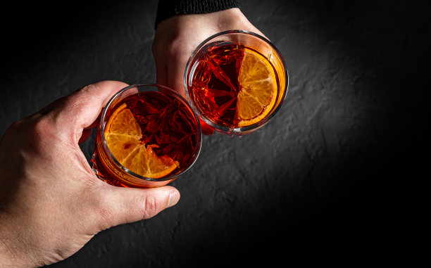 在黑色的背景下，男人和女人的手与内格罗尼鸡尾酒碰杯的特写。图片下载