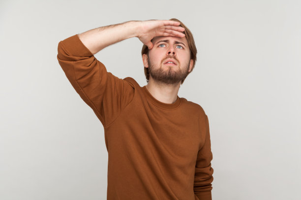 一个留着胡子穿运动衫的男人，手放在额头上站着找东西。图片下载