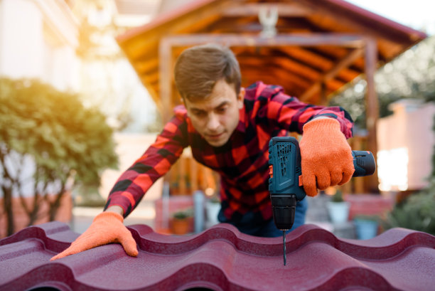 一个穿着格子衬衫，戴着防护手套的年轻人正在用电钻在金属瓦屋顶上钻孔。图片下载