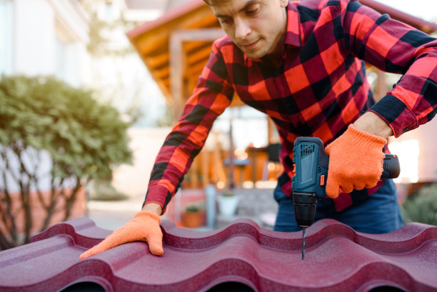 一个穿着格子衬衫，戴着防护手套的年轻人正在用电钻在金属瓦屋顶上钻孔。图片下载