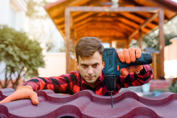 一个穿着格子衬衫，戴着防护手套的年轻人正在用电钻在金属瓦屋顶上钻孔。图片下载