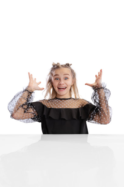 近距离拍摄漂亮可爱的小女孩，穿着黑色连衣裙的小学生坐在桌子旁，孤立在白色的工作室背景上。教育、童年的概念。图片下载