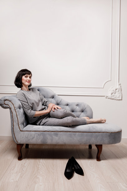 积极的年轻黑发女性光着脚坐在灰色沙发上的肖像，双腿伸展在灰色格子西装看着相机。女商人下班后脱鞋休息。图片下载