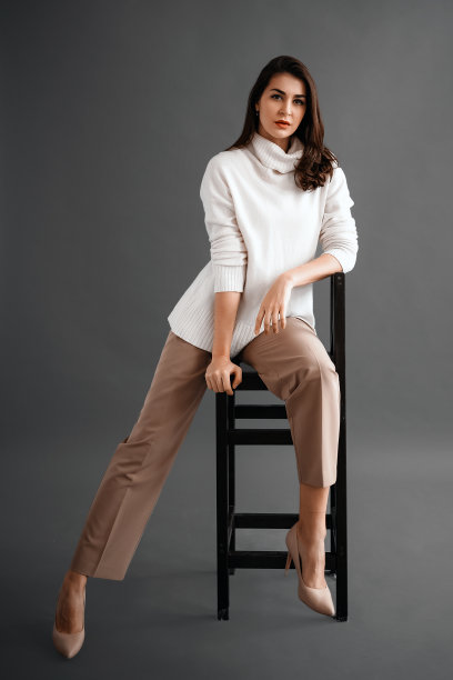 无忧无虑的年轻女商人坐在椅子上，穿着白色的毛衣和米色的裤子，在工作室的灰色背景下摆姿势。轻松而美丽的年轻女子图片下载