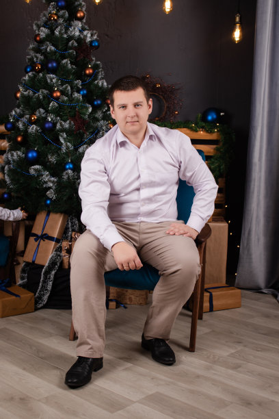 圣诞树和新年装饰品旁，一个穿着白衬衫和棕色裤子的英俊男子的画像。商务风格，办公室着装。商人图片下载