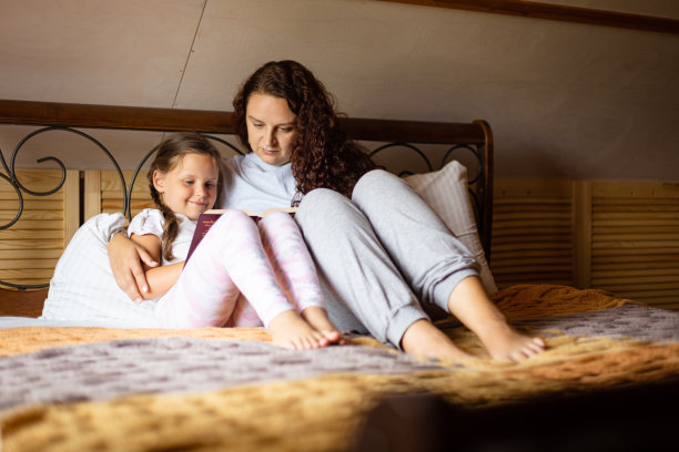 白天穿着家常服，和妈妈一起捧着书，坐在木屋里的双人床上，幸福的白人小女孩。家人在一起的时间图片下载