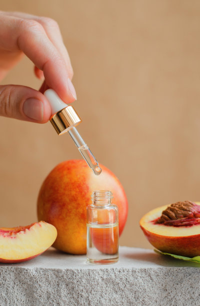 透明的瓶子与桃子油和新鲜的桃子在灰色的讲台上。自然皮肤和头发护理。一个女人手里拿着蘸着蜜桃油的滴管。天然的化妆品图片下载