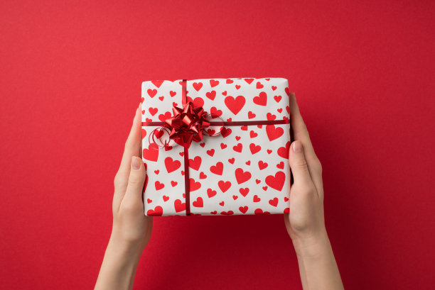 第一人称俯视图照片的情人节装饰年轻女孩的手拿礼品盒白色包装纸与心脏图案和红色的星弓在孤立的红色背景与copyspace图片下载