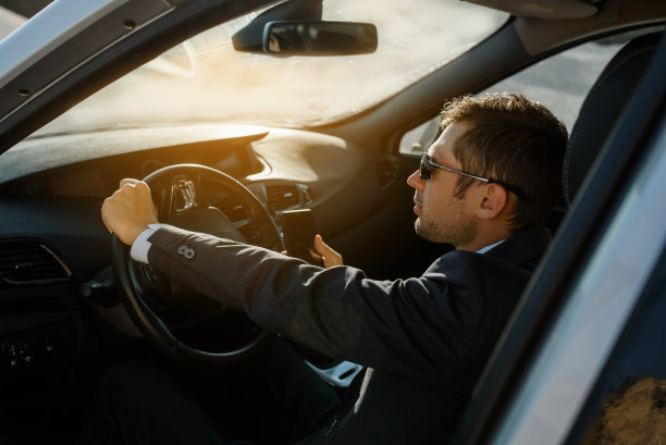 一位商人穿着夹克，衬衫，打着领带，戴着太阳镜，一边开车一边看他的手机。图片下载