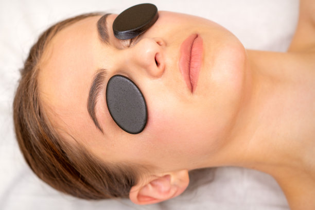 年轻女子在水疗中心接受眼部黑色矿物石的面部按摩。图片下载