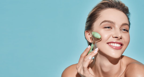 美丽的女人按摩她的脸用绿色的玉辊为弹性完美的皮肤，孤立在蓝色的背景与文字的空间。面部玉辊图片下载