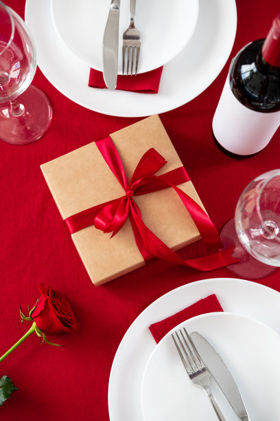 情人节的餐桌上摆放着餐具、礼品盒、酒瓶、玻璃杯、红玫瑰在红色的背景上。图片下载