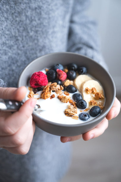 厨房里，一个身穿灰色毛衣的年轻女孩端着一碗素食酸奶、格兰诺拉麦片、新鲜的蓝莓和香蕉。健康早餐的概念图片下载