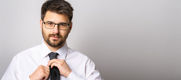 这是一名留着胡子、戴着眼镜的30岁白人成年男子的肖像，他穿着衬衫、打着领带，在白色的墙壁背景前看着摄像机，树立了事业自信和成功理念图片下载