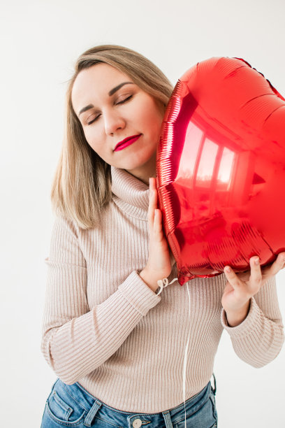 迷人的女孩闭着眼睛，抱着一个红色的心形气球微笑。情人节快乐图片下载