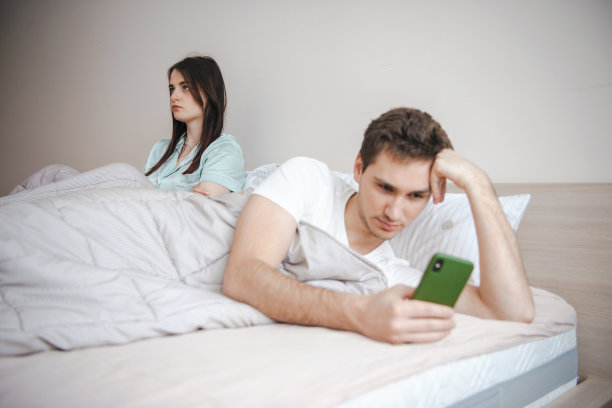 夫妻俩躺在床上，年轻的妻子坐着感觉心烦意乱，丈夫在午睡时间用智能手机图片下载