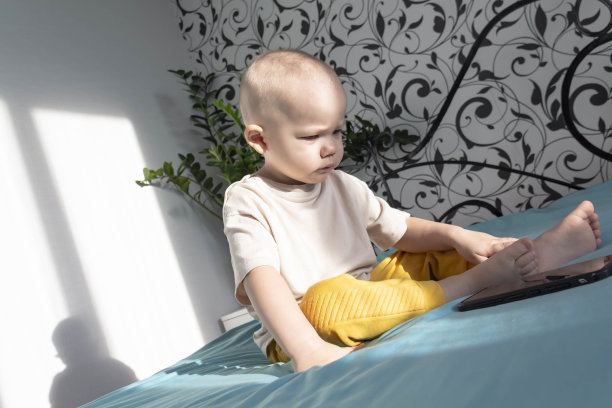 一个小男孩坐在床上的蓝色毯子上玩一部现代手机。图片下载