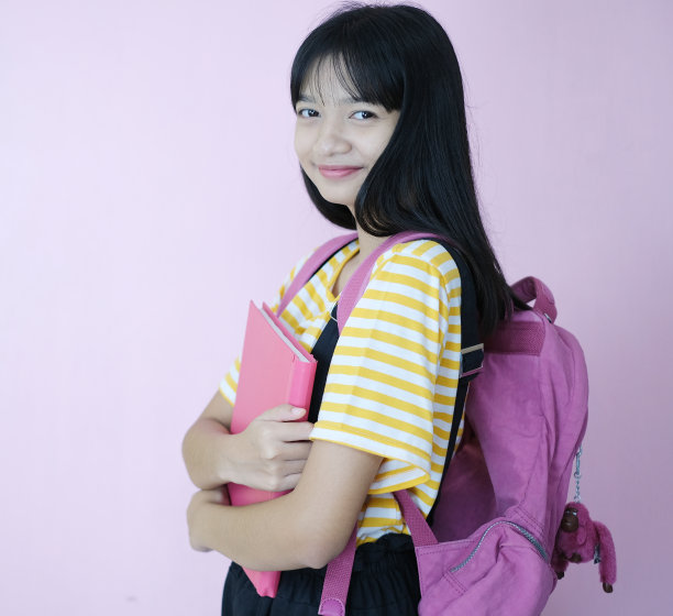 快乐的年轻学生拿着粉红色的书和粉红色背景的背包。图片下载