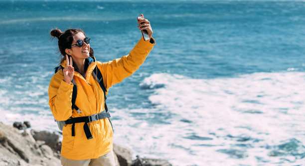 一个旅行者拿着手机站在大海的背景上。一名手持手机的女性游客，背景是海景。快乐的女游客在大海的背景上自拍图片下载