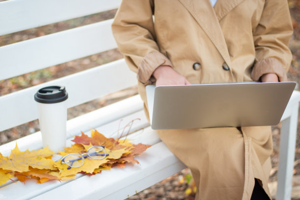 在公园里，一名穆斯林妇女戴着头巾，穿着米色风衣，端着一杯咖啡，坐在长椅上用笔记本电脑打字。学生。眼镜的景象。图片下载
