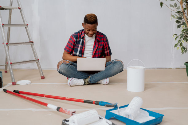 一名肤色黝黑的非裔美国人跪在公寓的纸箱上，手里拿着笔记本电脑，寻找灵感图片下载