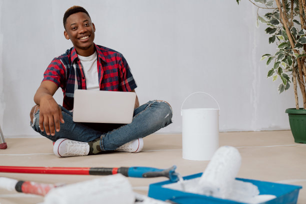 一名深色皮肤的男子正把笔记本电脑放在大腿上，坐在装修公寓的房间里寻找家具的灵感，他旁边躺着滚轮罐的白色油漆家伙微笑着对装修感到满意图片下载