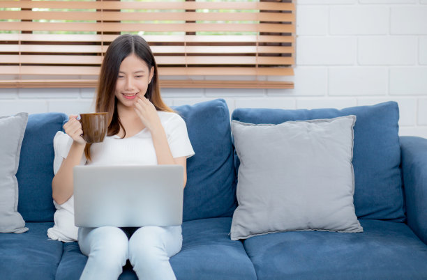 年轻的亚洲商业女性微笑着在家里用笔记本电脑在客厅的沙发上在线工作，自由职业女孩使用笔记本电脑和在沙发上喝咖啡，新常态，生活方式的概念。图片下载