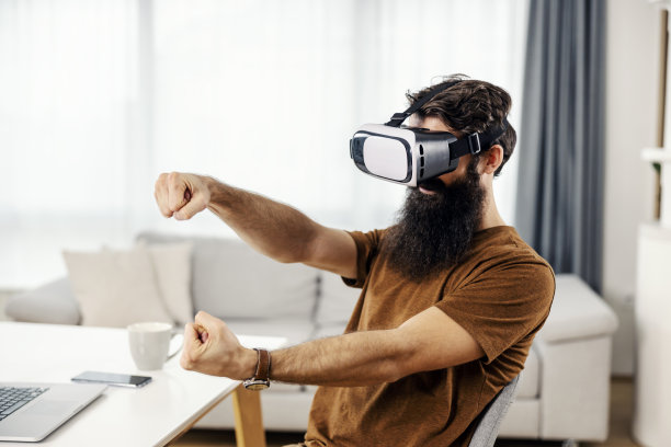 一个大胡子男人坐在他舒适的家里玩vr游戏，戴着vr头盔。图片下载