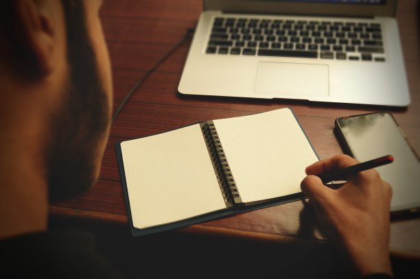 这张照片拍摄于一个年轻人的背部，他在一张空白的螺旋形笔记本上写字，在一张木制的桌子上用笔记本电脑和手机工作图片下载