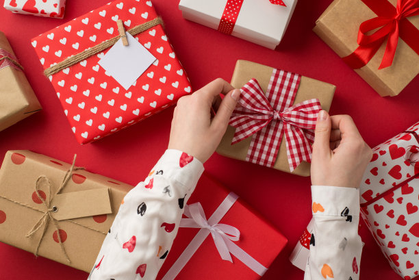 第一人称俯视图照片的情人节装饰礼物和女人的手在白衬衫系格子丝带蝴蝶结在孤立的红色背景的牛皮纸礼品盒图片下载