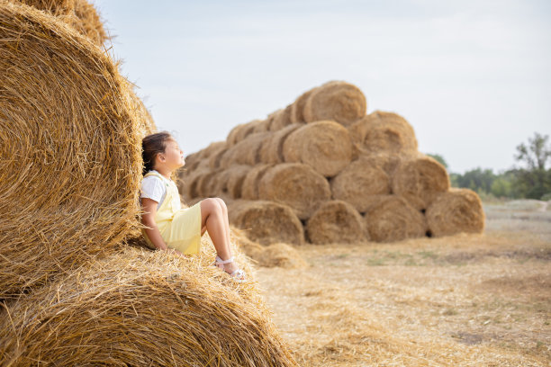 轻松的年轻女孩坐在高高的干草堆上，穿着太阳裙，靠在另一个干草堆上，闭着眼睛，抬头朝向天空。在满是金色干草的田野里享受远离城市的乐趣图片下载
