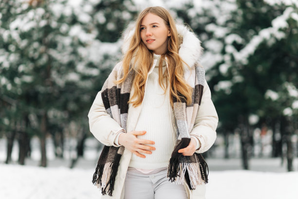 一名孕妇穿着暖和的冬衣，在一个下雪的冬天站在外面，双手捂着肚子图片下载