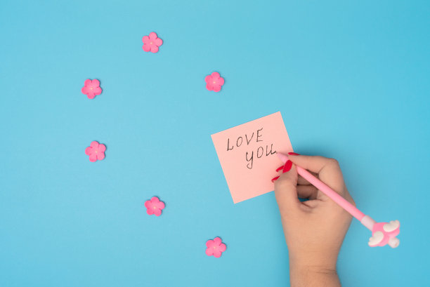 女性在粉红色的卡片便条上写下“爱你”，蓝色的背景上放着粉红色的小花。贺卡。爱情，情人节或其他节日的概念。平面布局风格与复制空间图片下载