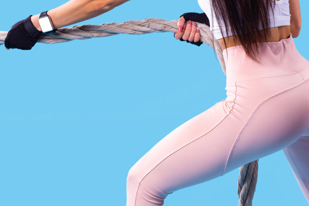 这是一个年轻苗条的女孩的臀部后视镜照片，她长着一头长发，穿着粉色的运动打底裤和白色的上衣，正在用健身绳锻炼，她正在把绳子拉向自己。图片下载