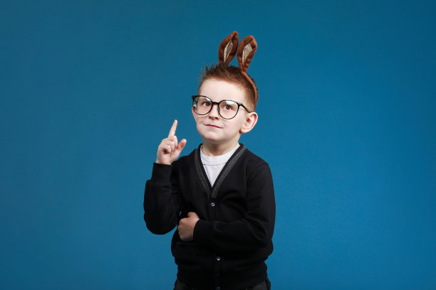 复活节快乐的孩子。小男孩戴着兔子耳朵，戴着眼镜，在蓝色画室背景上。指向上。看着镜头微笑。图片下载