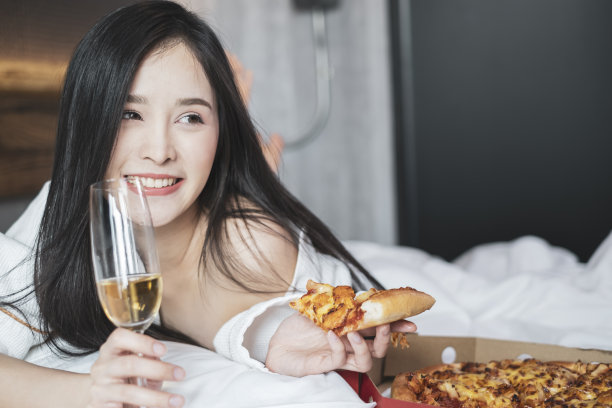 快乐的年轻女子在酒店房间的床上吃披萨喝香槟。图片下载