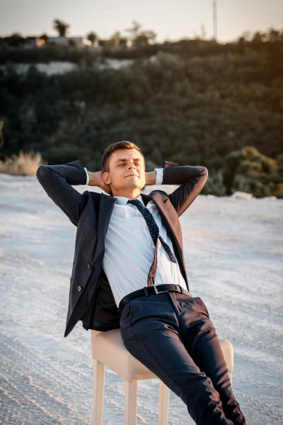 一个年轻人穿着一件经典的夹克，双手放在脑后，坐在椅子上休息。图片下载