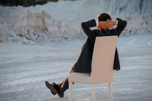 一个年轻人穿着一件经典的夹克，手放在脑后，坐在椅子上休息。图片下载