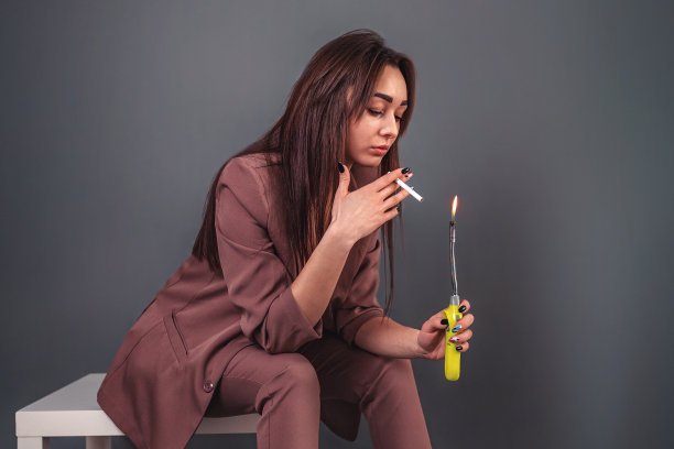 尼古丁上瘾的概念。白种年轻女子，穿着西装，坐在凳子上，点着一支烟。灰色的背景。近距离图片下载