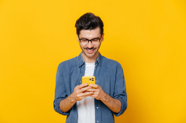 一个帅气的白人男子正用他的智能手机，和朋友发短信，浏览互联网和社交网络，站在一个孤立的橙色背景上，微笑着图片下载