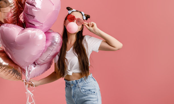 年轻女子跳舞，微笑着，在一根粉色心形气球的大树枝旁玩泡泡糖。孤立的粉红色背景。图片下载