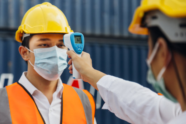 亚洲男性工人戴着口罩，在施工现场为预防新冠肺炎、新冠肺炎或冠状病毒大流行而进入工作之前，用数字体温计检查发烧情况。有选择性的重点。图片下载