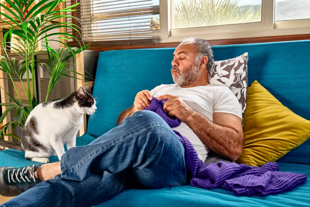 一名留着胡子的中年男子坐在沙发上，用毛线球编织针编织一条紫罗兰色的羊毛围巾，一边和他的猫玩耍，一边编织，这是他保持积极心理健康的爱好之一图片下载