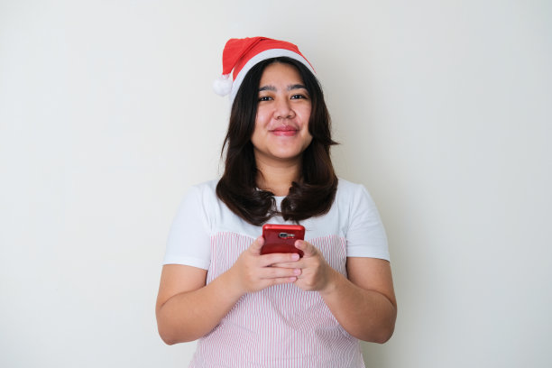 成年亚洲妇女戴着圣诞帽微笑，而持有手机图片下载