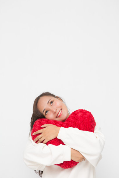 美丽的亚洲妇女穿着白色毛衣拥抱红色心形枕头的白色背景和复制空间的情人节概念。图片下载